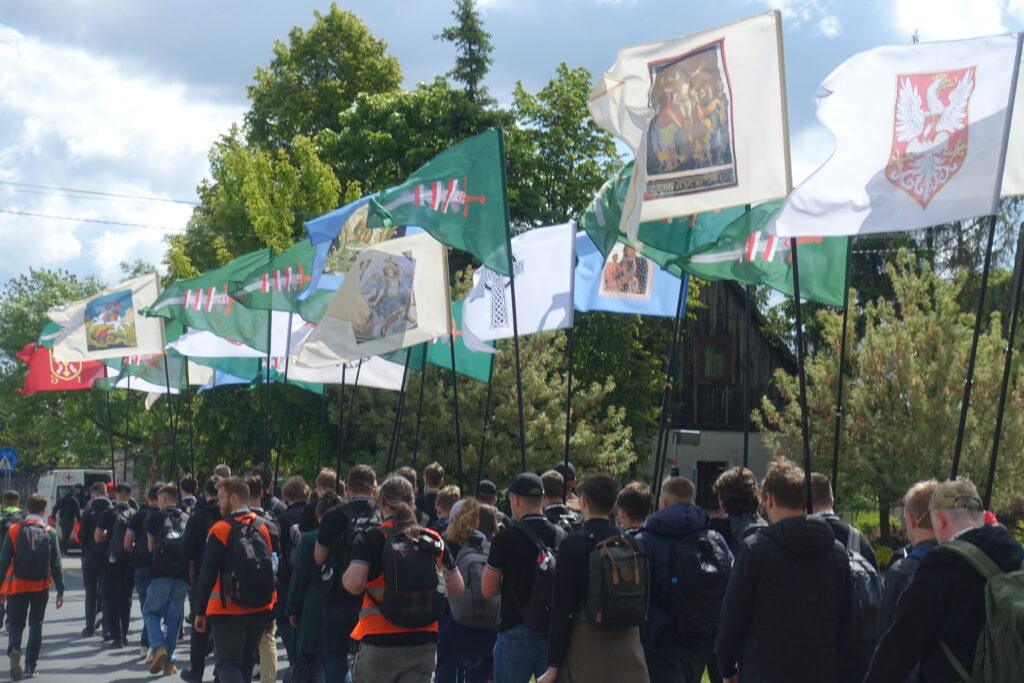 Grupa pielgrzymów Pielgrzymki Kościoła Walczącego maszeruje z chorągwiami przedstawiającymi świętych i symbole narodowe.