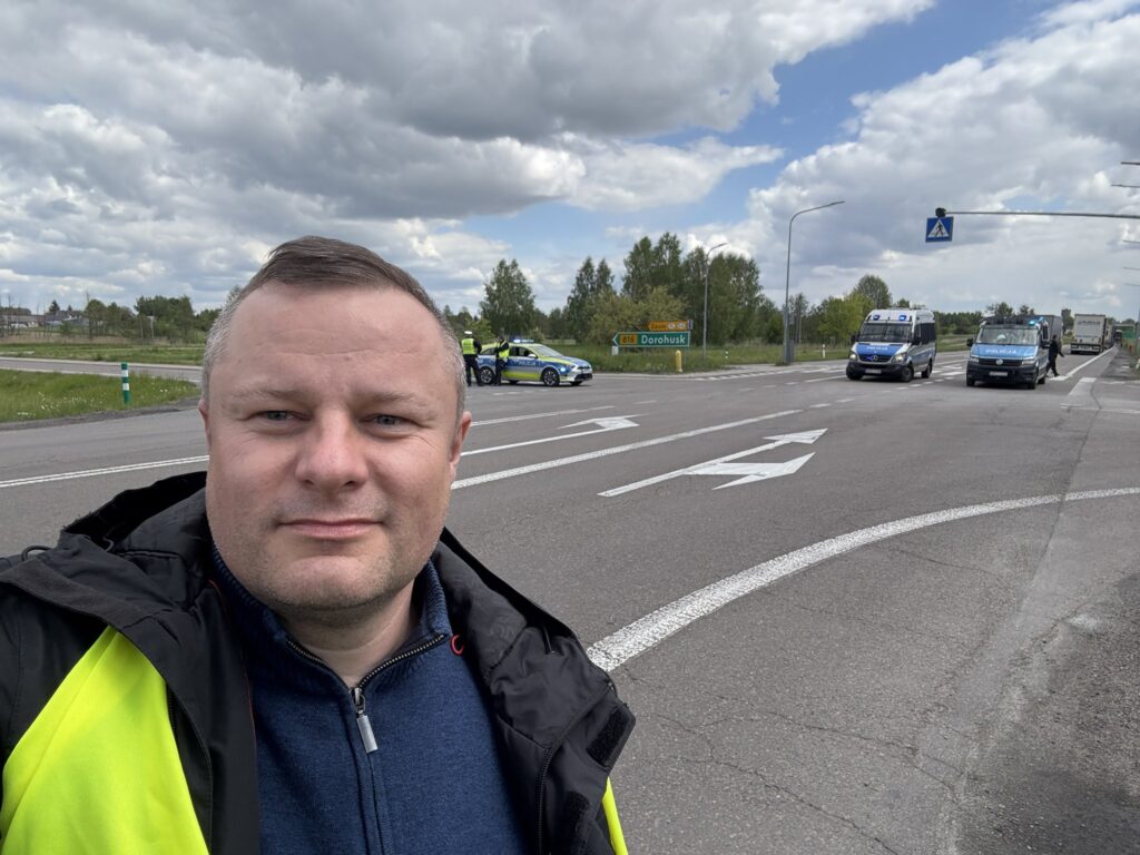 Rafał Mekler na tle blokady drogowej w Dorohusku, z widocznym oznaczeniem drogi 816 i obecnością policji.